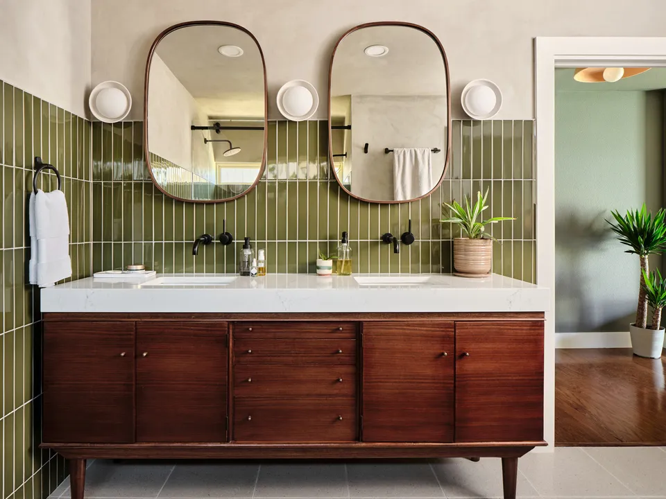 Mid-century walnut double vanity below two oval mirrors and green vertical tile