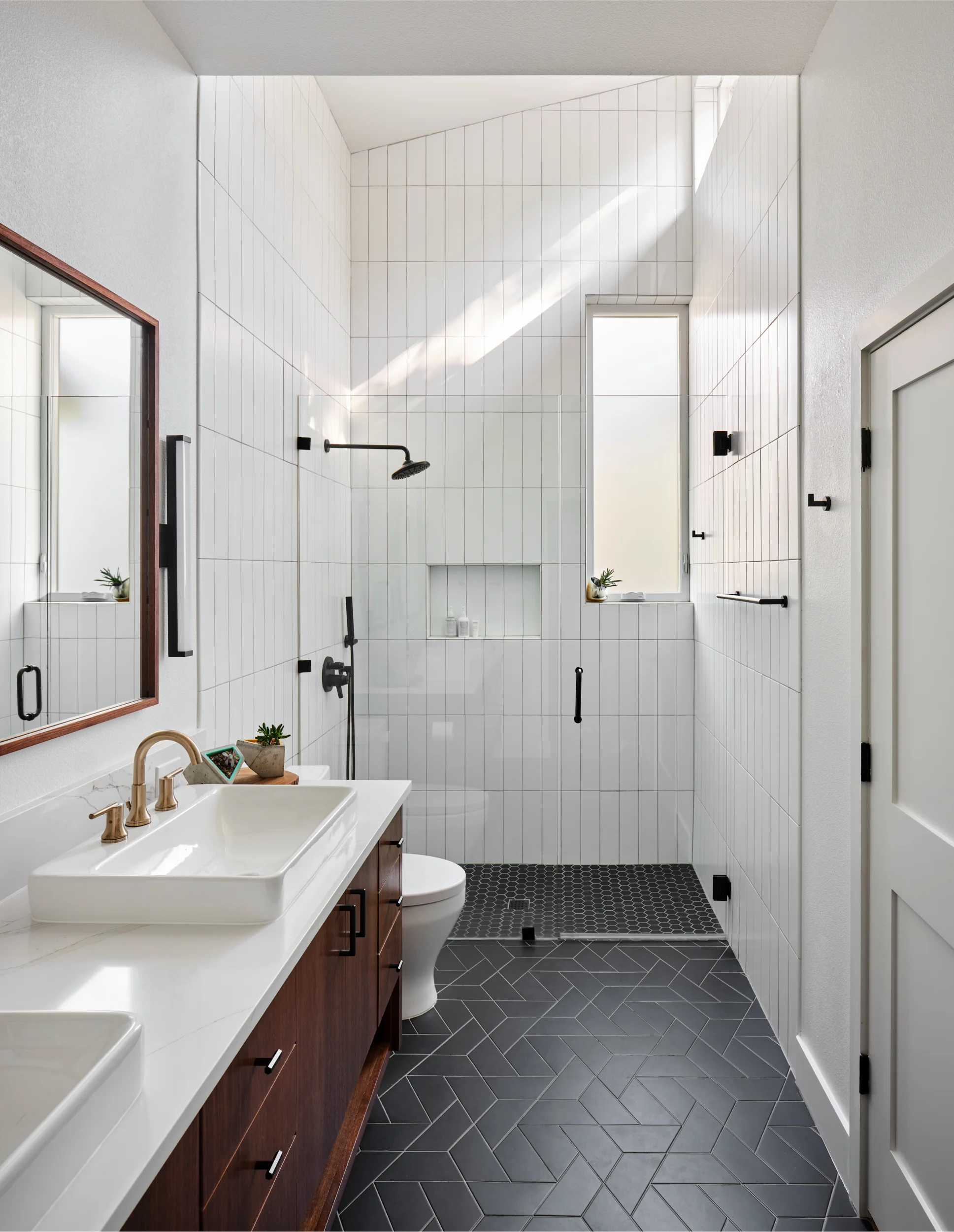 Walk-in shower with white vertical tile and black fixtures beside a wood vanity with a brass faucet