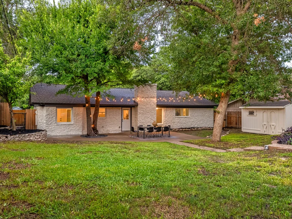 Back lawn under trees with the white stone house and string lights