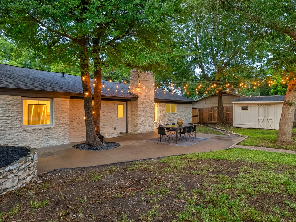Backyard patio with string lights under a tree beside the white stone house