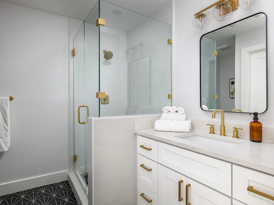 Glass shower beside a white vanity with brass hardware and a black-framed mirror