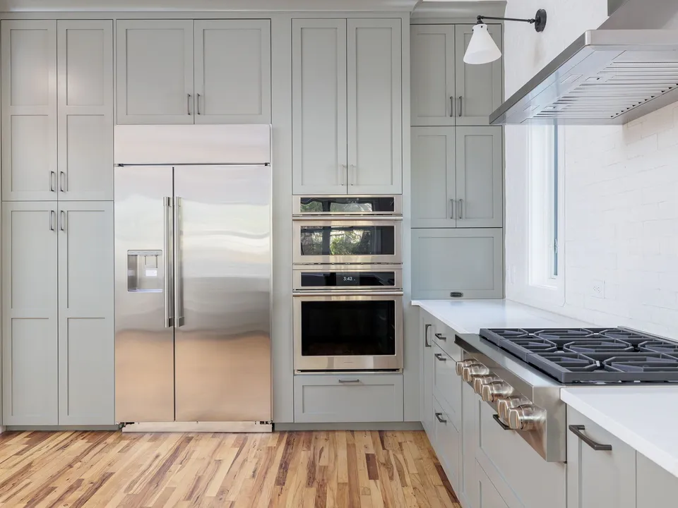 Built-in stainless refrigerator and double wall ovens in grey shaker cabinets beside a gas cooktop