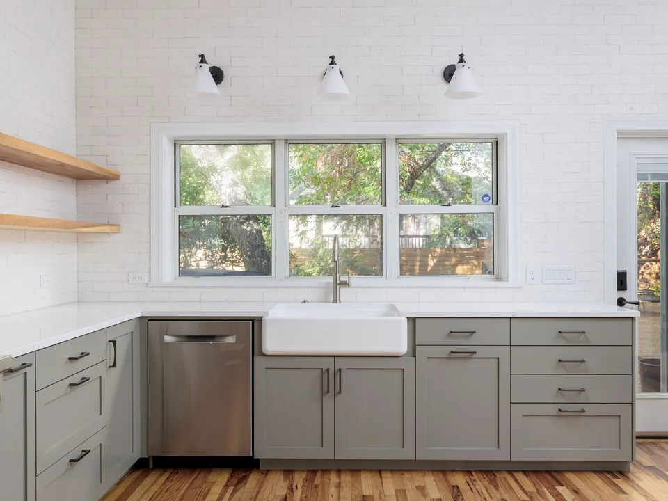 Farmhouse sink below a window in grey shaker cabinets with open wood shelves