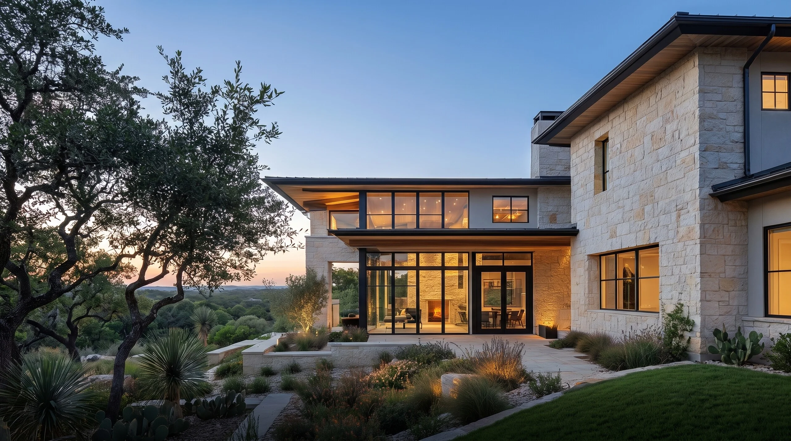 Two-story limestone home at dusk with a glass-walled great room, lit windows and native landscaping under live oaks