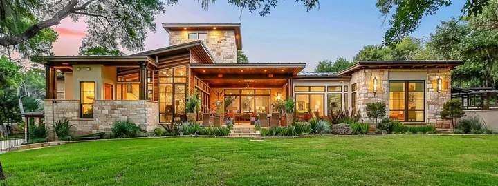 Limestone home with a covered wood-ceiling patio and lit windows at dusk, behind a wide lawn under live oaks