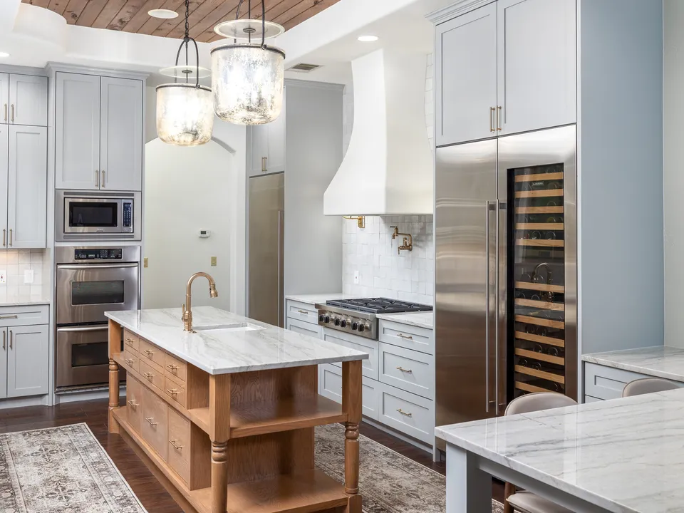Stain-grade island below lantern pendants, with double ovens and a wine fridge beyond