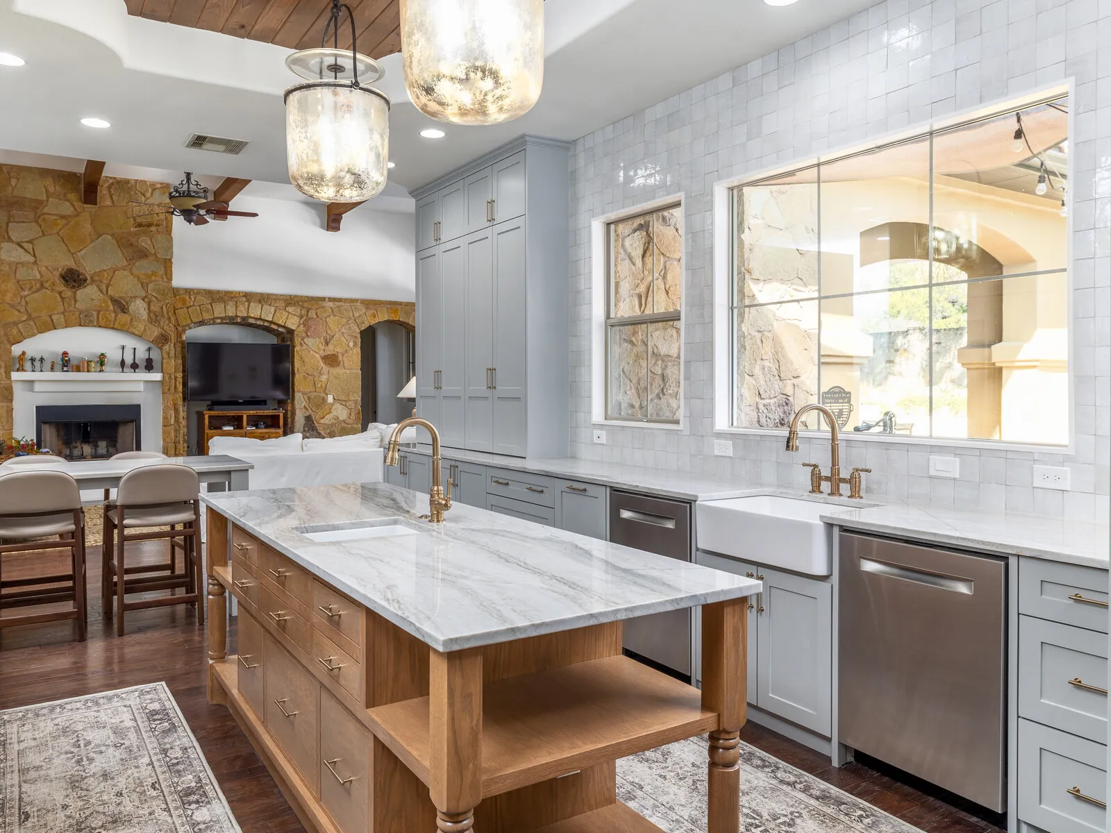 Kitchen island with a wood base, turned legs and open shelves under glass lantern pendants, with a stone fireplace wall beyond