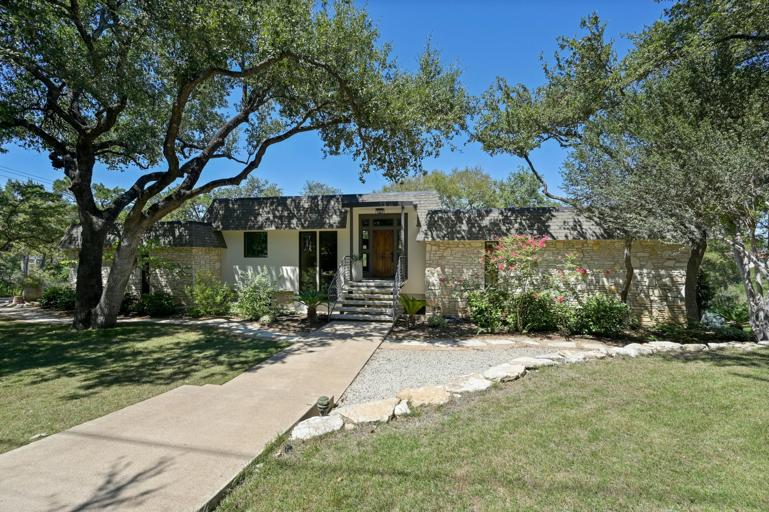 Single-story stone and stucco house with a wood front door and entry steps, set among large oak trees behind a front walk