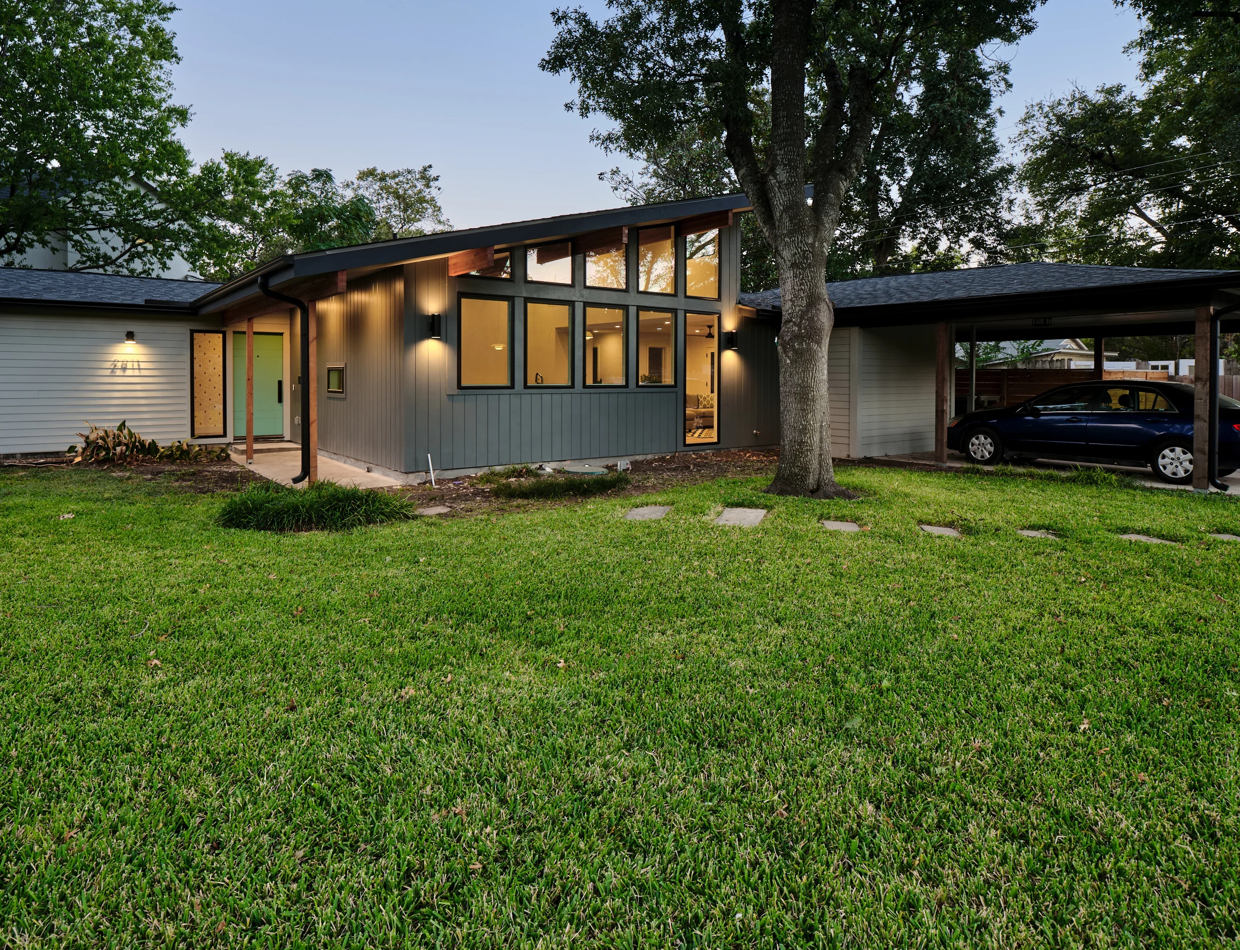 Mid-century modern house at dusk with a single-pitch roof, lit clerestory windows and dark board-and-batten siding beside a carport