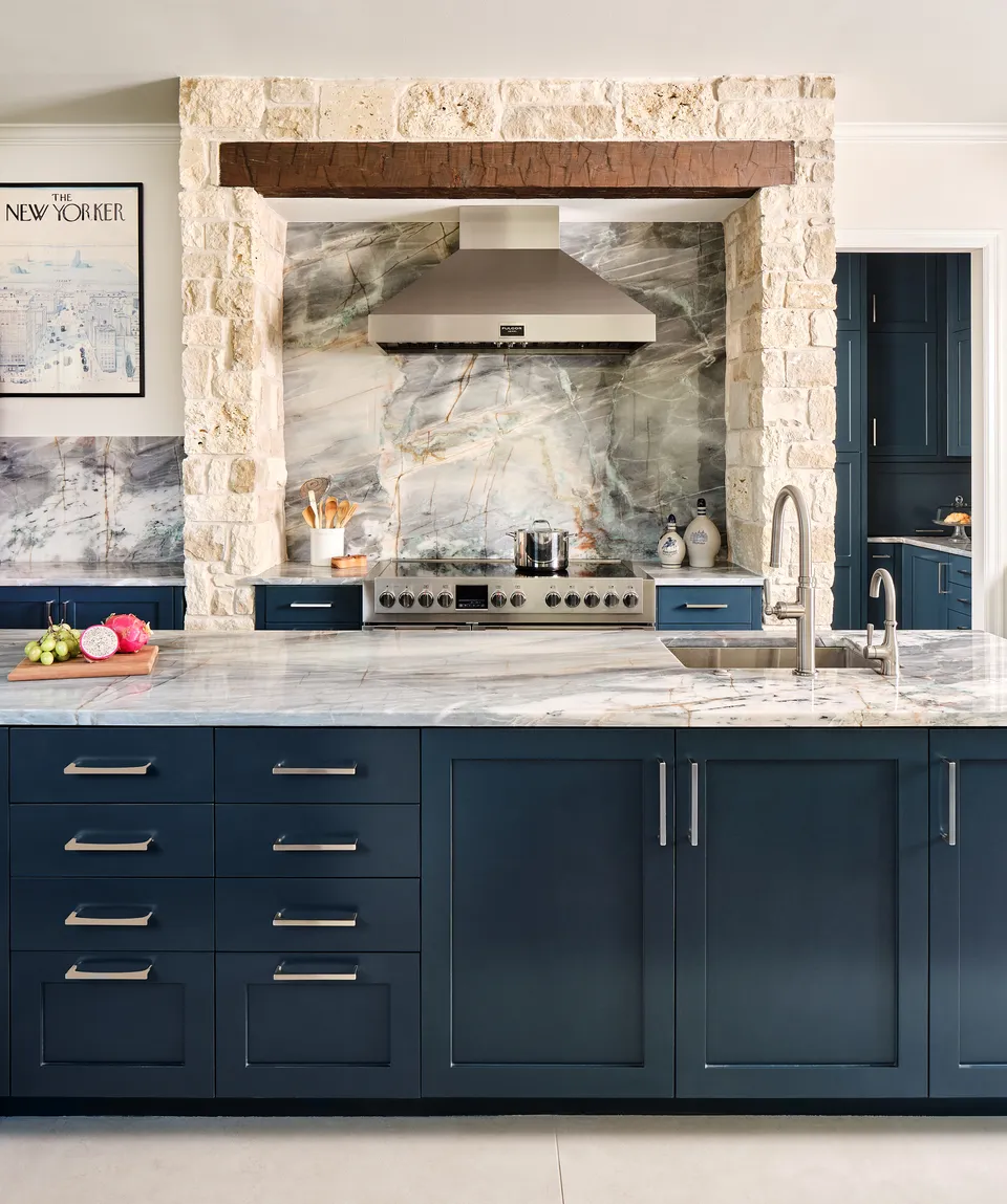 Navy kitchen cabinets and a range under a stainless hood, set in a stone surround with a wood beam and a veined slab backsplash