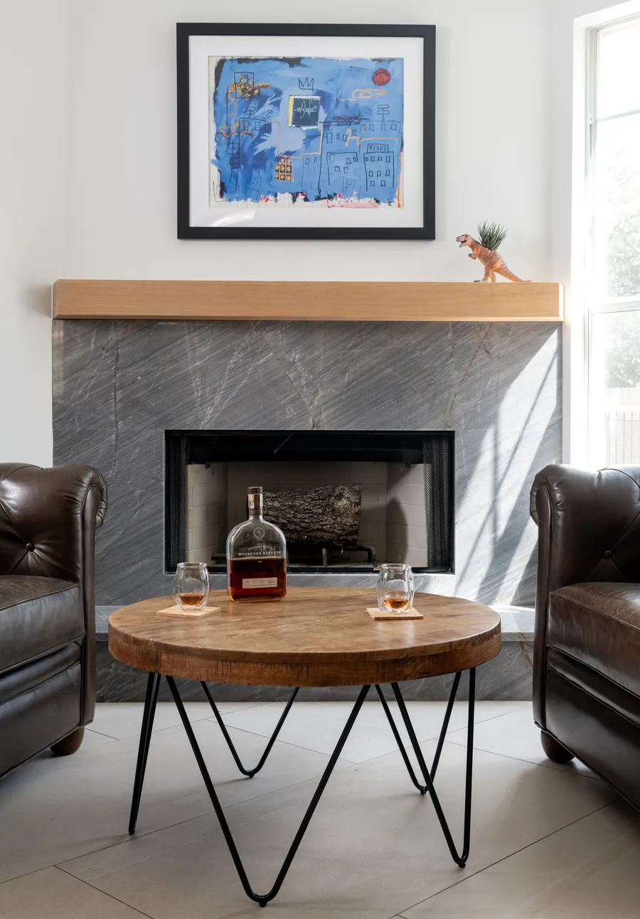 Fireplace with a grey stone surround and a wood mantel between leather chairs and tall windows