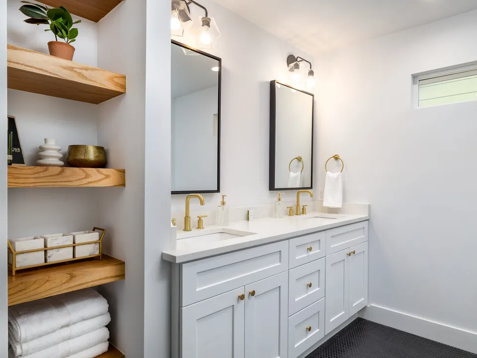 Primary bath with wood open shelves, a white double vanity and brass fixtures
