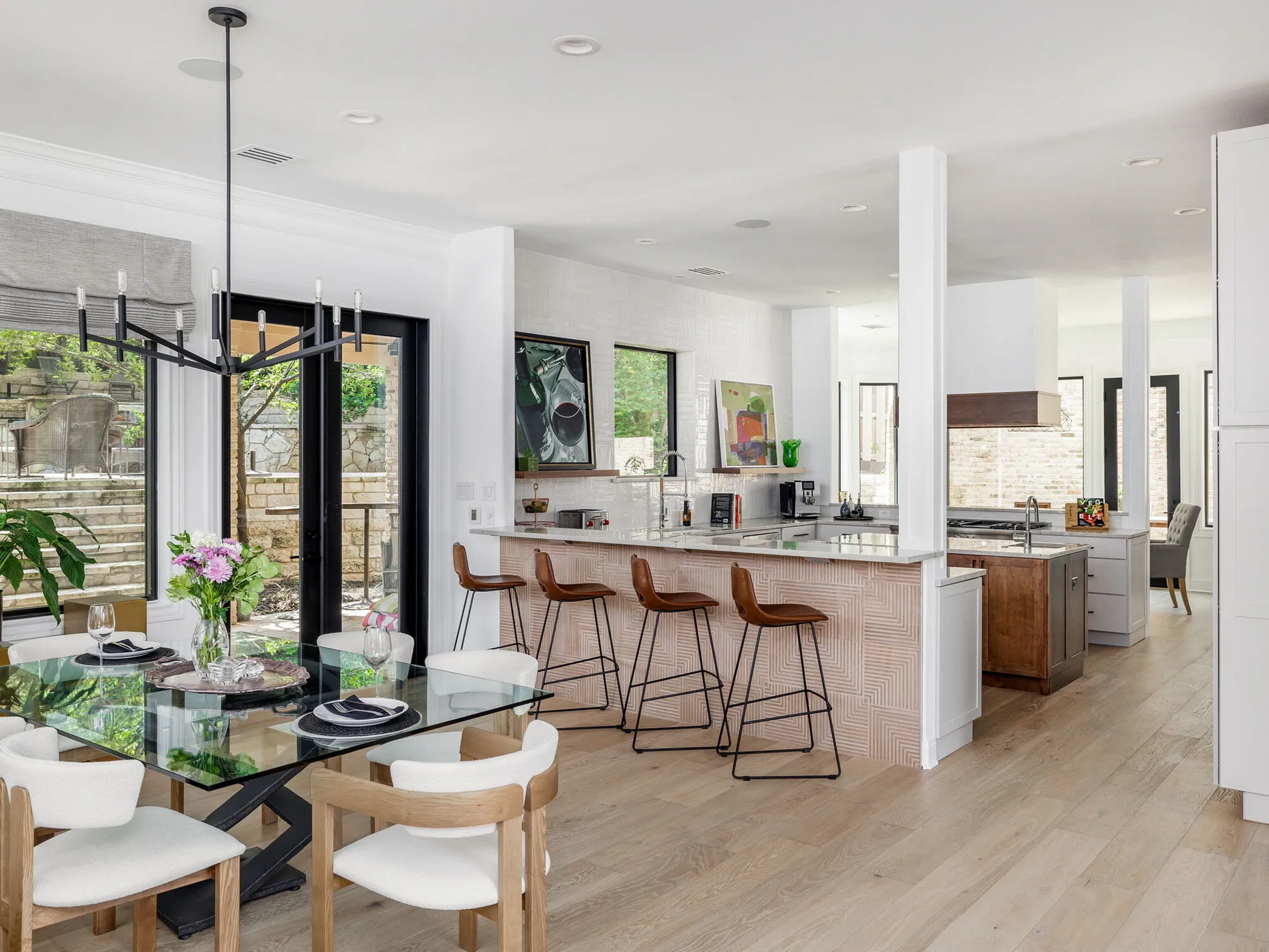 Dining area with a glass table and white upholstered chairs open to a kitchen peninsula with leather bar stools