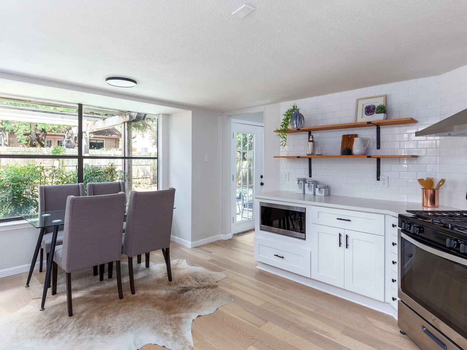 Dining area with a glass table in a window bay beside a white kitchen