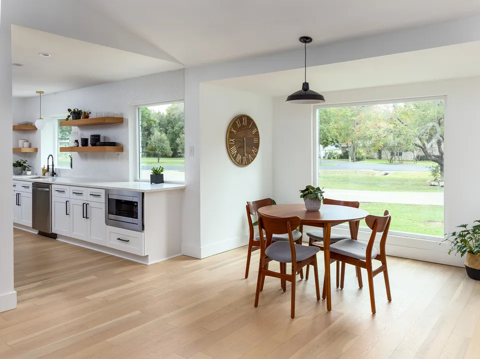 Breakfast nook with a round table below large windows, beside a white kitchen