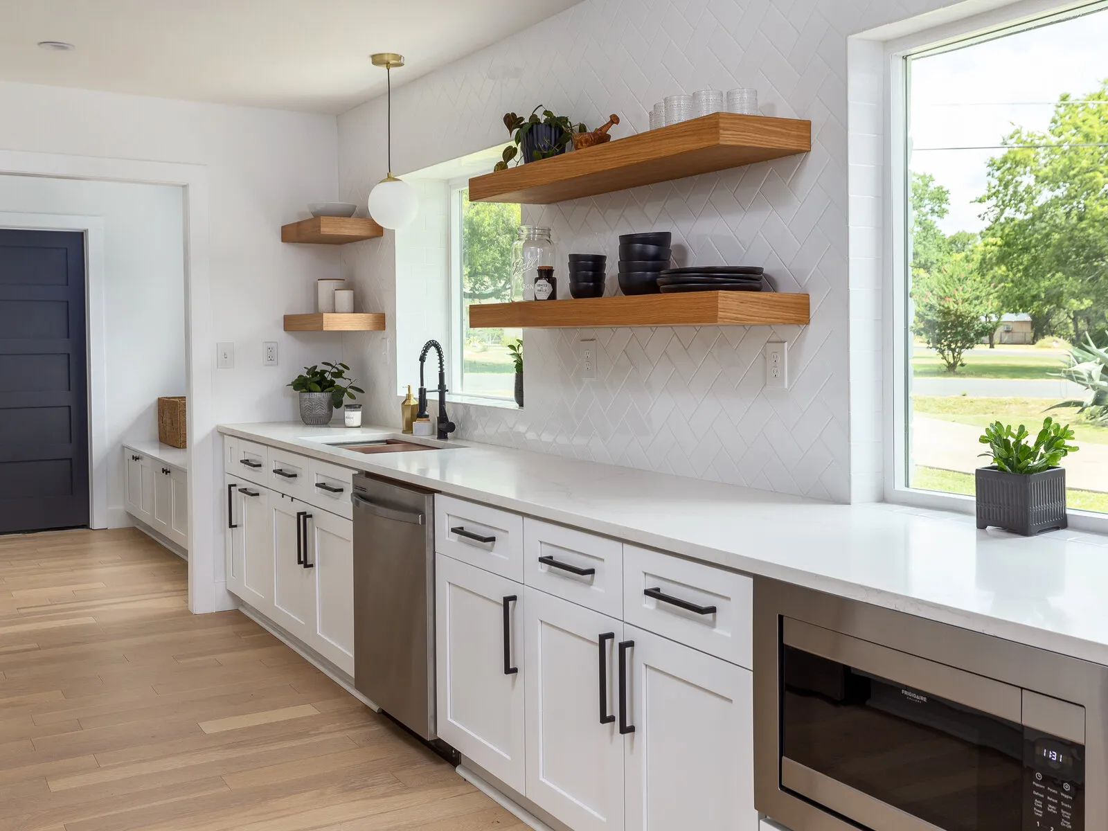 White shaker kitchen with walnut floating shelves beside a large window and a stainless dishwasher