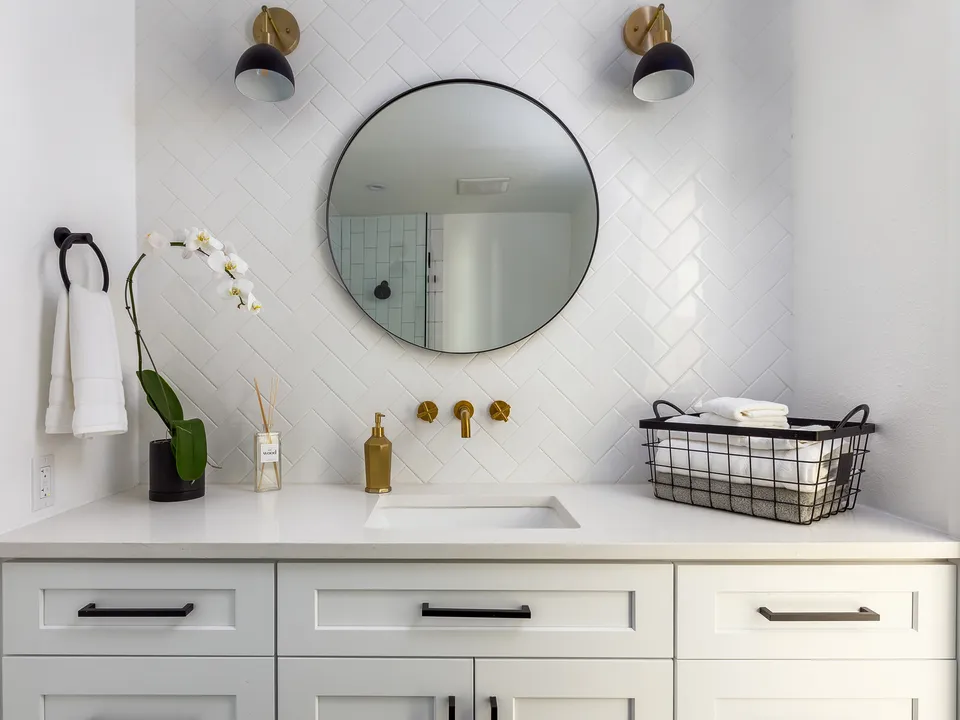 White vanity with a round mirror, black sconces and white herringbone tile