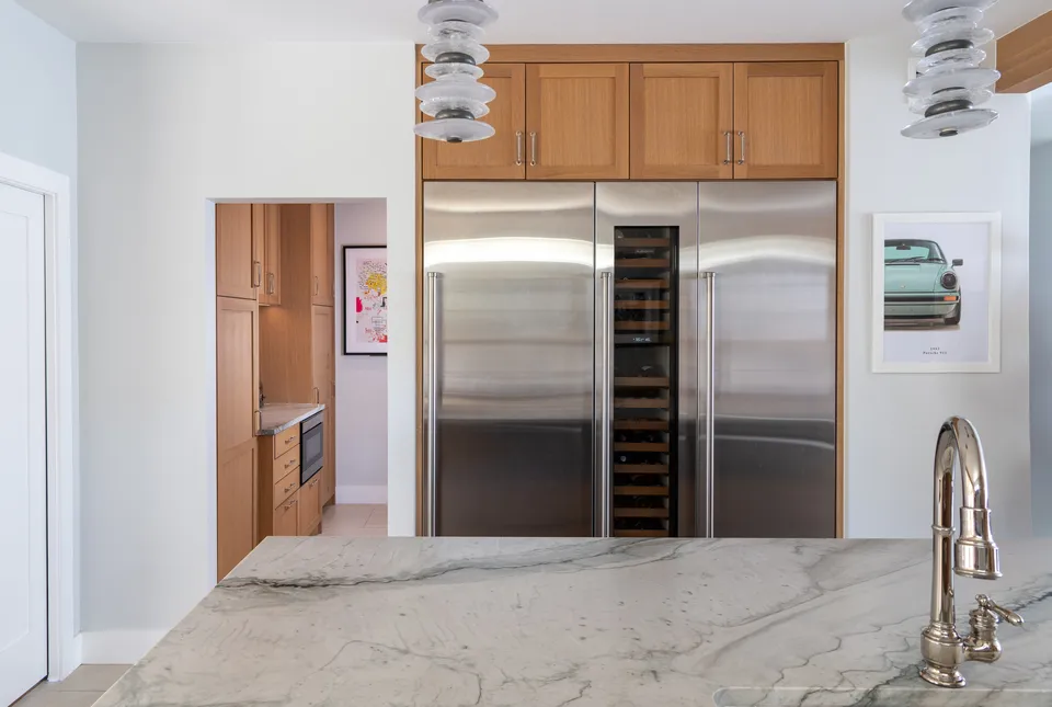 Built-in stainless refrigerator and wine column in white oak cabinetry, seen across a marble counter