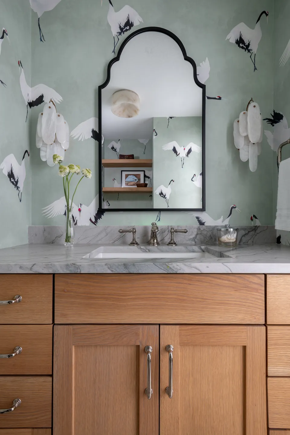Oak vanity with a marble top below an arched black mirror on crane wallpaper