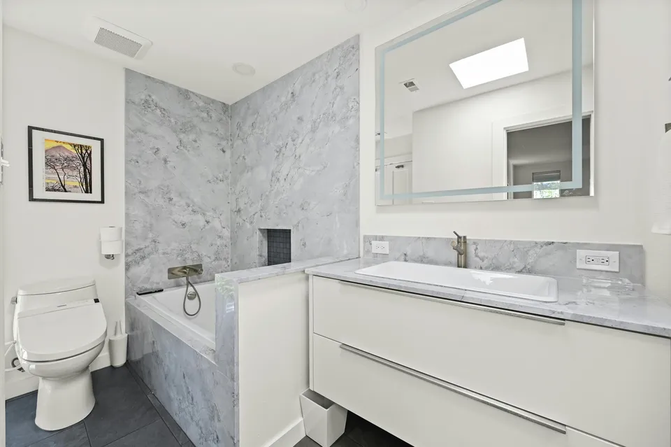 Bathroom with a marble tub surround and a lit mirror over a white vanity