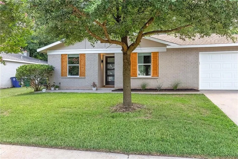 Front of the painted brick house with orange shutters behind a large tree