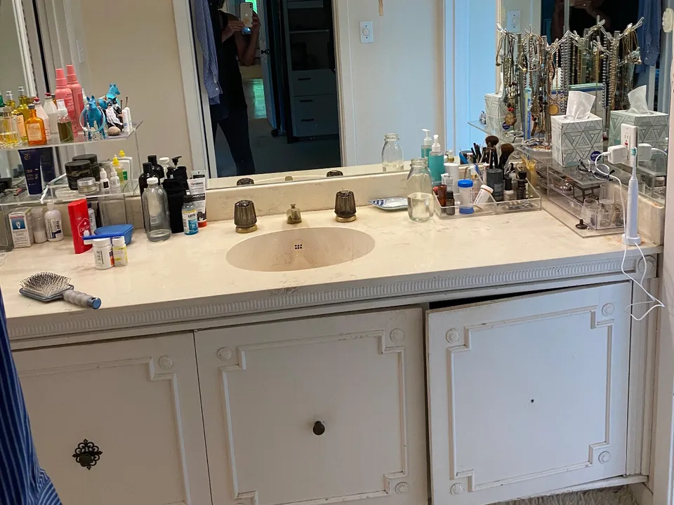 Bathroom before the remodel, with a cluttered white vanity and dated cabinet doors