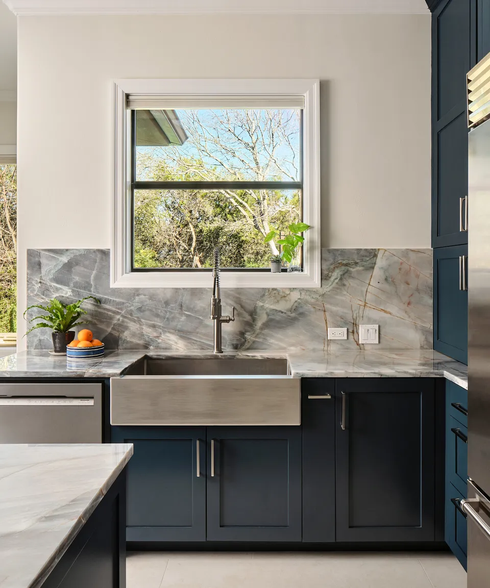 Stainless farmhouse sink below a window with a quartzite slab backsplash and navy cabinets