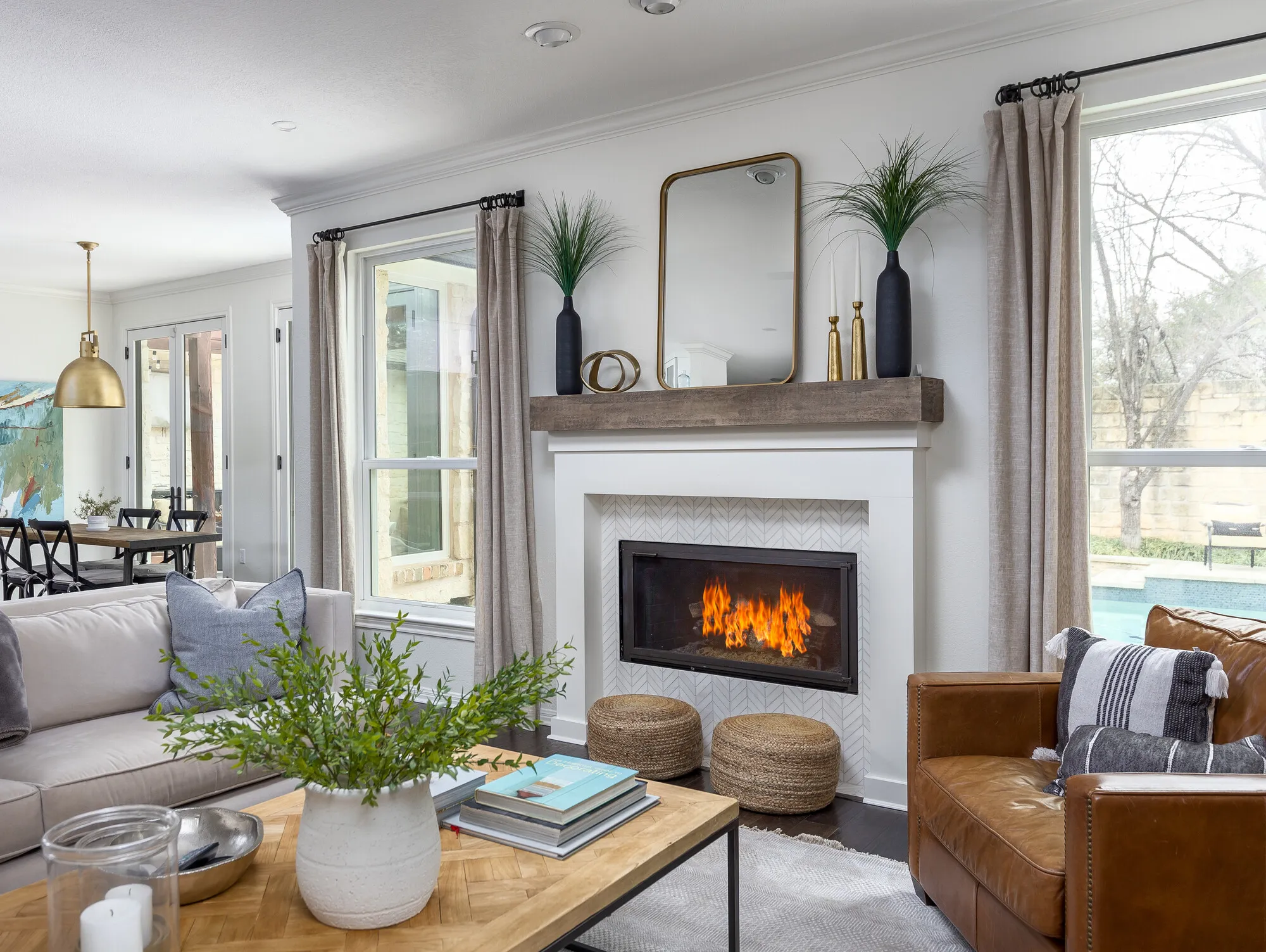 Fireplace with a white herringbone tile surround and a wood beam mantel under a brass mirror, between two tall windows