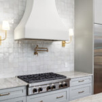 beautifully remodeled kitchen with upscale stainless steel cooktop and fridge. One side of the fridge is full of wine drawers. Above the cooktop there are two brushed brass sconce lights and a brushed brass pot filler. Above that you will find a large custom plaster hood vent. hand made tile backsplash is off white and a natural stone countertop.