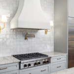 beautifully remodeled kitchen with upscale stainless steel cooktop and fridge. One side of the fridge is full of wine drawers. Above the cooktop there are two brushed brass sconce lights and a brushed brass pot filler. Above that you will find a large custom plaster hood vent. hand made tile backsplash is off white and a natural stone countertop.