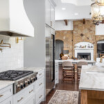 Austin kitchen remodeler's newly remodeled kitchen with grey shaker cabinets. The backsplash is made from square tiles that are hand made and white with a grey hint of color in them. complete with brass fixture including a pot filled over the cooktop and sconce lights surrounding the cooktop. Stainless Steele fridge and cooktop with a white custom made range hood built from white plaster